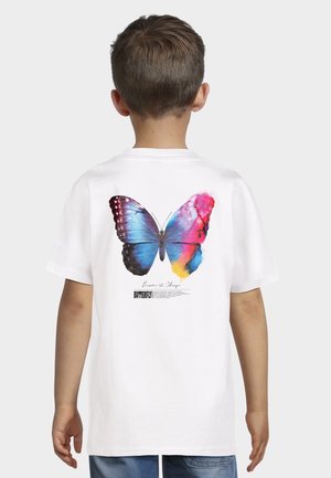 Dziecko ubrane w białą koszulkę z dużym kolorowym motylem i napisem „Zostań zmianą” na plecach, stojące na białym tle.