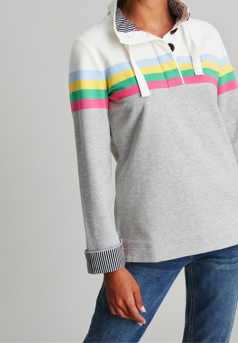 Femme portant un sweat à capuche à manches longues avec des rayures bleues, jaunes, vertes et roses sur un tissu blanc et gris clair, assorti à un jean bleu.