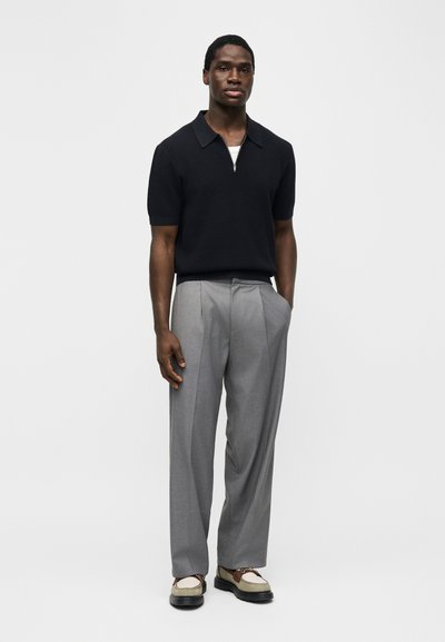 Άνδρας που στέκεται φορώντας μαύρο κοντομάνικο πόλο με φερμουάρ, ανοιχτό γκρι πλισέ παντελόνι και μπεζ δετά παπούτσια με μαύρες σόλες.