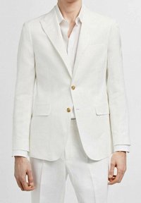 Blazer bianco in lino con due bottoni dorati, rever a punta e tasche laterali, indossato sopra una camicia leggera. Tessuto liscio, vestibilità sartoriale.