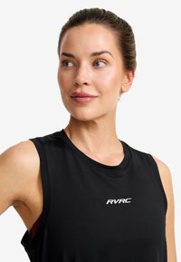Woman with pulled-back hair wearing a black sleeveless top with "RVRC" logo, looking slightly to her left against a plain background.