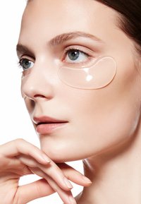 Des patchs en gel transparent placés sous les yeux d'une femme à la peau claire, au teint lisse et à la couleur des lèvres neutre. Un éclairage doux met en valeur ses traits.
