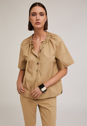 Beige blouse met korte mouwen en gathered halslijn details, knoopsluiting en een ontspannen pasvorm, gecombineerd met bijpassende broek en een geometrische armband.