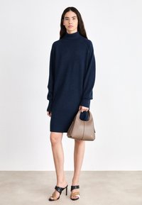 Nő tengerészkék, hosszú ujjú pulóverruhában, taupe színű kézitáskával, fekete és barna színű magassarkú szandálokkal, egyszínű háttér előtt áll.