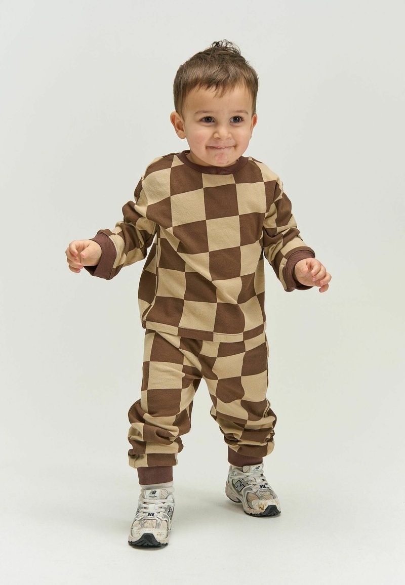 Tout-petit debout, portant un sweat-shirt et un pantalon à carreaux beige et marron, avec des baskets blanches, sur un fond clair uni.