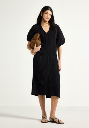 Kvinde står iført en sort tekstureret midikjole og sorte sandaler, holder en brun blød clutchtaske med et lyst armbånd om håndleddet.