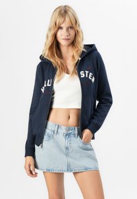 Young woman with long blonde hair wearing a navy zip-up hoodie, white crop top, and light denim skirt, posing with one hand in pocket.
