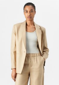 Blazer en lin beige avec des épaules structurées et une fermeture à un bouton, associé à un débardeur à côtes gris clair et un pantalon assorti.