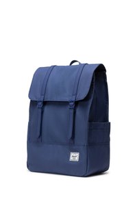Dunkelblauer Rucksack mit Klappe, zwei vorderen Riemen, seitlicher Tasche und einem Markenetikett. Hergestellt aus strapazierfähigem Stoff mit glatter Oberfläche.