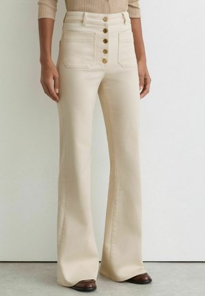 Pantalon évasé taille haute couleur crème avec cinq boutons dorés et poches avant, porté avec un haut beige côtelé à manches longues.