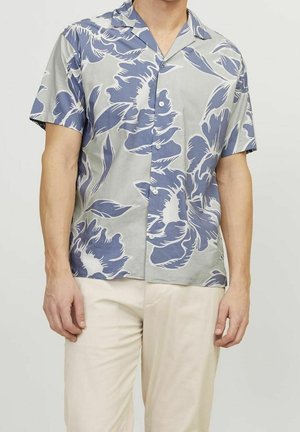 Mann trägt kurzärmliges Hemd mit großem blauem Blumenmuster und hellbeiger Hose, steht vor einfachem Hintergrund.