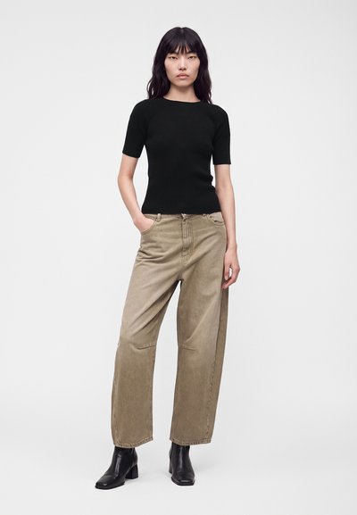 Femme portant un haut noir à manches courtes, un pantalon ample beige et des bottines noires, debout avec une main dans une poche sur fond blanc.