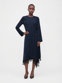 Femme portant une robe bleu marine à manches longues avec bordure en dentelle sur un ourlet asymétrique, debout avec une main sur la hanche et portant des talons foncés.