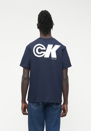 Mand med dreadlocks iført en mørkeblå Calvin Klein T-shirt med stort hvidt "CK"-logo på ryggen og blå jeans, stående mod en ensfarvet baggrund.