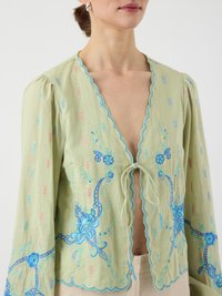 Kvinne i lys grønn, brodert bluse med blå blomsterdesign, bølget kant, puffermer og knytelukking foran.