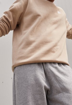 Person trägt beige Nike-Sweatshirt und graue Jogginghose, mit leicht erhobenen Armen vor neutralem Hintergrund.