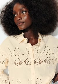 Blouse beige clair avec des motifs de broderie découpée élaborés, un col et une fermeture à boutons, en tissu doux et texturé.