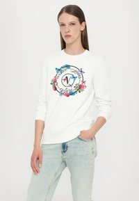 Felpa bianca con una grafica floreale rotonda, testo in nero e farfalle blu, abbinata a jeans in denim blu chiaro. Materiale in cotone morbido.