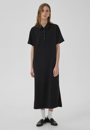 Kobieta ubrana w czarną midi sukienkę z krótkim rękawem, zapinaną na guziki przy kołnierzyku, z białymi skarpetkami i czarnymi mokasynami, stojąca na tle jednolitego tła.