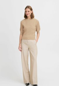 Maglione beige lavorato a maglia con design a trecce testurizzate e maniche a puff corte, abbinato a pantaloni a gamba larga beige chiaro e scarpe nere.