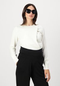 Pull blanc à manches longues avec un design texturé, associé à un pantalon noir taille haute. Le mannequin porte de grandes lunettes de soleil noires et des boucles d'oreilles argentées en forme de cercle.