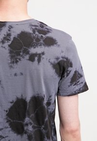 Šedé bavlněné tričko s černým tie-dye vzorem. Design obsahuje vířící tvary a textury, má kulatý výstřih a krátké rukávy.
