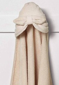 Beige handduk med texturerad tyg, som har en huva och en dekorativ rosett. Mjuk, absorberande material, hängande mot en neutral bakgrund.