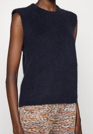 Haut sans manches en maille duveteuse bleu marine foncé porté avec un pantalon texturé multicolore marron et blanc sur une personne.