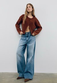Cardigan côtelé couleur rouille avec un design court, associé à un jean large bleu clair, terminé par des talons aiguilles marron.