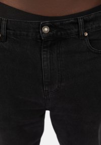 Czarne dżinsy z przodem kieszeniami, metalowym zapięciem na guzik i gładkim pasem. Tkanina ma lekki połysk i dopasowany krój.