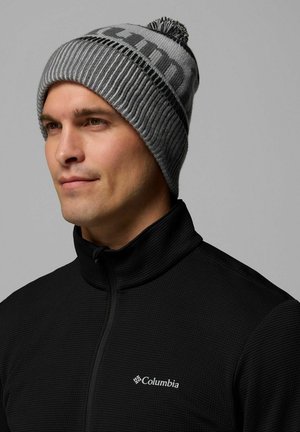 Berretto di maglia grigio con texture a coste, caratterizzato da una fascia nera con la scritta "Columbia" e un pompon sulla parte superiore. Indossato con un colletto nera con zip.
