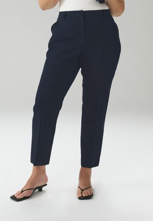 Naine, kes kannab kohandatud tumesiniseid pahkluuni pükse ja musta värvi madala kontsaga sandaale, seisab ühevärvilise heleda tausta ees.