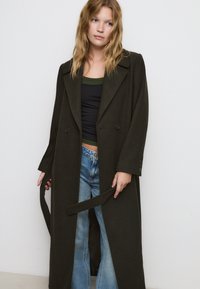 Cappotto lungo di colore verde scuro, realizzato in una misto lana, con colletto a revers, chiusura a doppio petto con bottoni e fiocco laterale. Indossato sopra una maglietta nera e jeans.