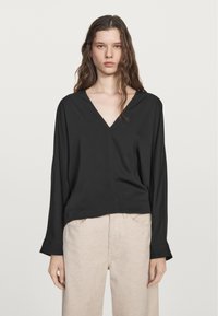 Blusa nera a maniche lunghe con scollatura a V, in tessuto morbido, vestibilità rilassata e orlo dritto, abbinata a pantaloni chiari e testurizzati.