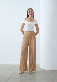 Haut blanc décolleté avec un détail de devant torsadé, associé à un pantalon large plissé doré. Tissu lisse et sandales neutres.
