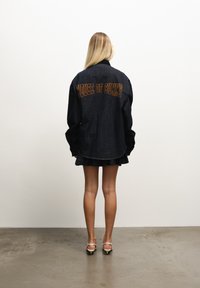 Dongerijakke i mørkeblått med brodert "HOUSE OF SUNNY" i oransje på ryggen. Har overdimensjonerte ermer og buet kant.