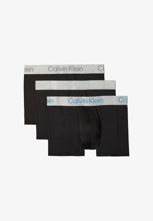 Muskulöser Mann steht barfuß mit verschränkten Armen, trägt schwarze Calvin Klein Boxershorts vor einem schlichten grauen Hintergrund.