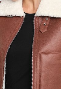 Giacca in pelle marrone con colletto in shearling bianco, zip metallica e tasche laterali. Superficie in pelle testurizzata e vestibilità su misura.