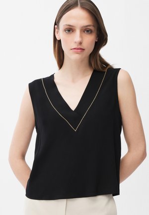 Jeune femme aux cheveux bruns raides portant un haut noir sans manches à col en V orné de passementerie dorée, debout devant un fond blanc uni.