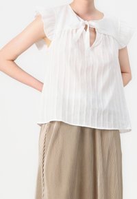 Weiß gestreiftes Oberteil mit Rüschensäumen und Schlüssel-Ausschnitt, ausgestattet mit einer gebundenen Schleife. Kombiniert mit einem beigen, plissierten Rock.