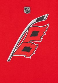 Camiseta roja con un palo de hockey y dos banderas rojas estilizadas con acentos negros. Logotipo de la NHL impreso encima del diseño gráfico.