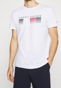 Camiseta de algodón blanca de manga corta, con un diseño de rayas negras y rojas y el texto "TOMMY HILFIGER" en el pecho.