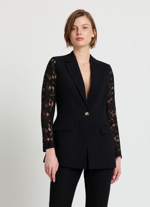 Woman with short hair wearing a black blazer with lace sleeves and matching lace pants, standing against a plain light background.