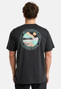 Black cotton t-shirt featuring a round graphic with wave and sun design in mint and peach colors, text reads "Billabong Adventure Division."