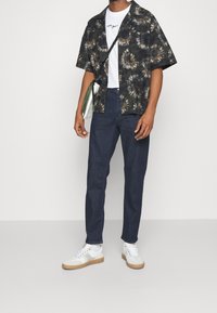 Maglietta nera a maniche corte con motivo floreale, t-shirt bianca, jeans scuri e sneakers bianche con suola beige; tiene in mano una borsa verde.