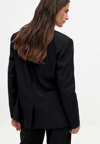 Black tailored blazer with a structured fit, featuring eyelet details on the sleeves and a single vent at the back. Smooth texture.