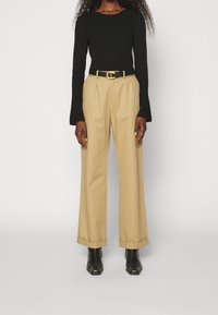 Haut à manches longues en maille côtelée noire associé à un pantalon beige taille haute à jambes larges, ceinturé d'une ceinture noire à boucle dorée. Bottines noires.