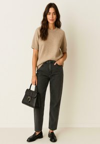 Vrouw in een beige trui met korte mouwen, hoge zwarte jeans, zwarte loafers, met een zwarte handtas, staande tegen een effen muur.