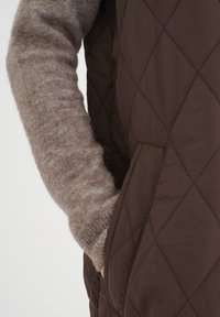 Quiltad brun väst med sidofickor, bärs över en ljusbrun, strukturerad tröja med ärmar. Fokusera på diamantmönstret och mjuka material.