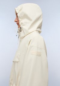 Giacca a vento beige chiaro con cappuccio realizzata in materiale resistente all'acqua. Presenta una tasca frontale, polsini elastici e una toppa con il marchio sulla manica.